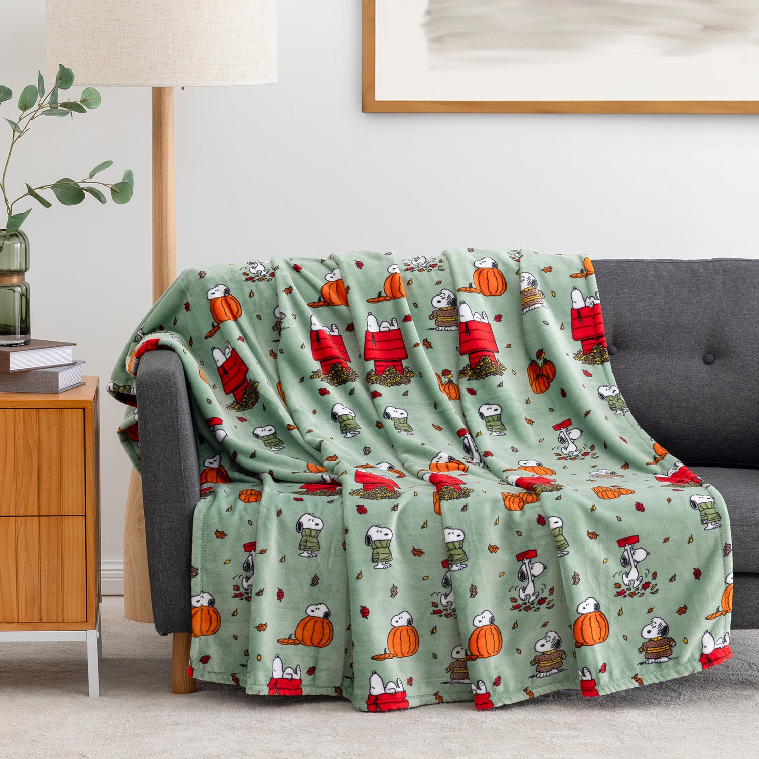 A sage green fleece throw blanket with a cartoon pumpkin and character print is draped over a gray sofa in a living room setting.