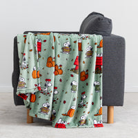 A mint green throw blanket with a repeating cartoon print of Snoopy and Woodstock in autumn outfits, draped over the back of a dark gray armchair.