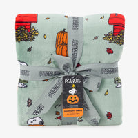 A folded Peanuts-themed throw blanket in a pale mint green color, decorated with autumn and holiday motifs including pumpkins, leaves, and red houses. The blanket is wrapped in a gray ribbon printed with 'PEANUTS' and has a hanging tag.
