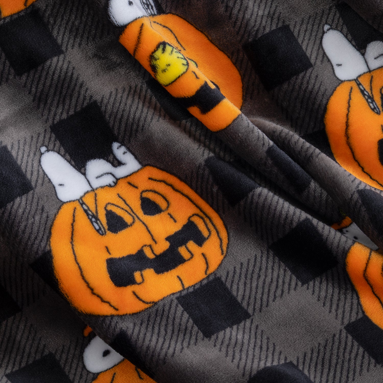 Close-up of a fabric surface with a Halloween pattern featuring orange jack-o'-lantern pumpkins, white cartoon dog faces, and black-and-gray check blocks.