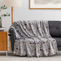A gray throw blanket with an all-over Snoopy cartoon print is draped over a dark gray sofa in a styled living room setting.