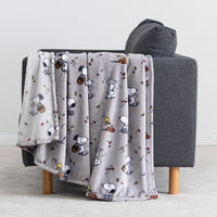 A light gray plush throw blanket with a repeating Snoopy and Woodstock autumn pattern, draped over a dark gray armchair with wooden legs in a clean indoor setting.