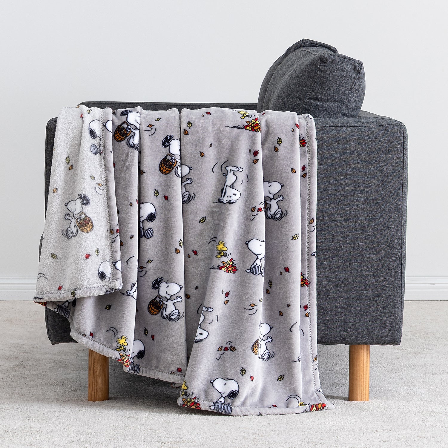 A light gray plush throw blanket with a repeating Snoopy and Woodstock autumn pattern, draped over a dark gray armchair with wooden legs in a clean indoor setting.