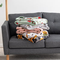 Four folded blankets with Halloween-themed prints are stacked on a dark gray fabric sofa. The blankets feature designs including a mint green pumpkin patch, a pink spider, a white pumpkin patch, and an orange-and-black tiger patch.