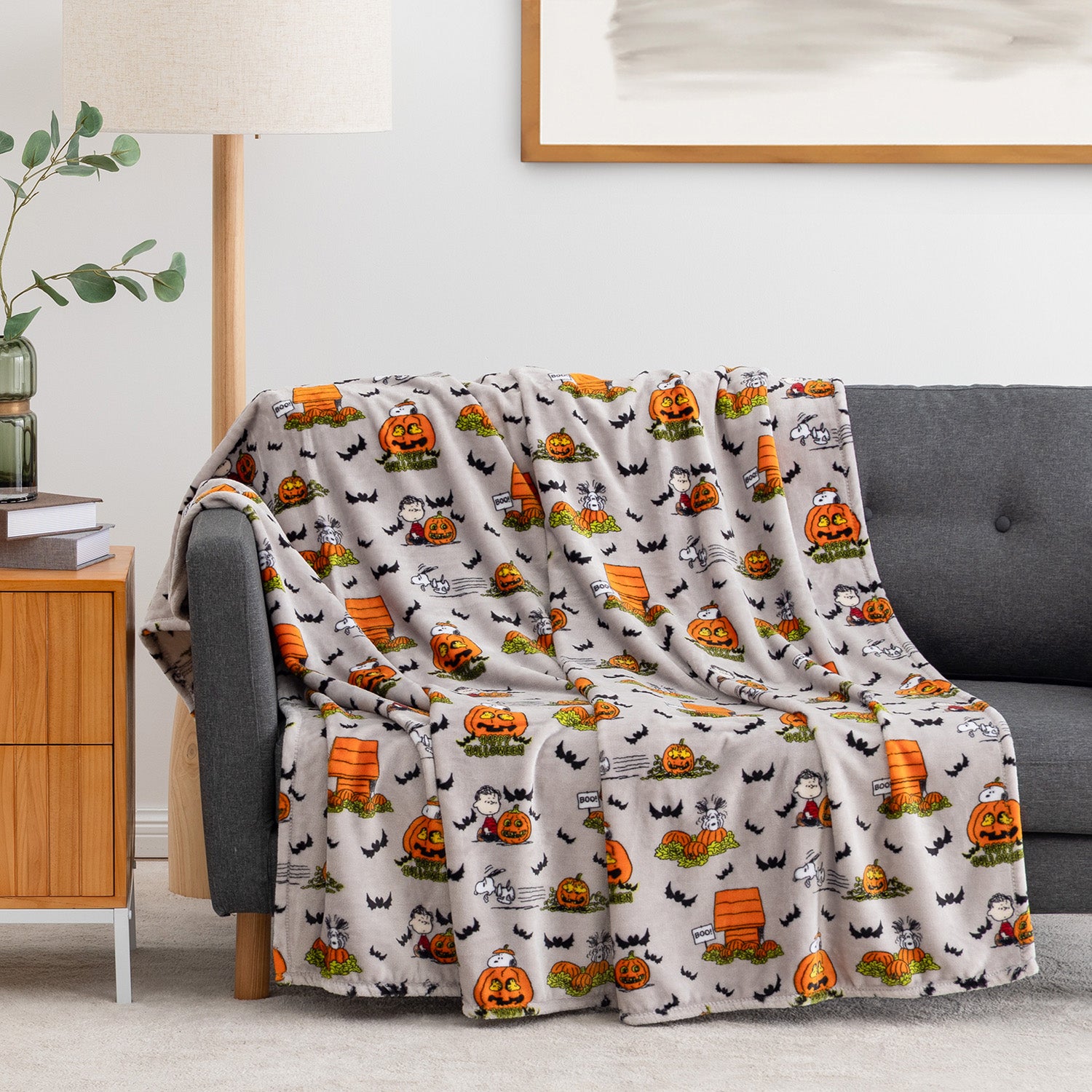 A fleece throw blanket with a Halloween pattern featuring pumpkins, bats, ghosts, and mummies, draped over a gray sofa in a living room setting.