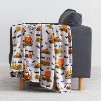 A fleece throw blanket with a Halloween-themed cartoon print featuring Snoopy and Charlie Brown, pumpkins, bats, and a haunted house, draped over a gray armchair.