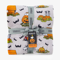 A folded plush throw blanket featuring a Peanuts Halloween print with Snoopy, Woodstock, pumpkins, bats, and a doghouse on a light gray background, wrapped with a gray ribbon and a Peanuts hangtag.