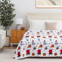 A bed dressed in a white comforter or sheet set printed with a repeating cartoon pattern of Snoopy and his doghouse, pumpkins, leaves, and autumn motifs, styled in a bright bedroom with a wooden nightstand, lamp, plant, and white headboard.