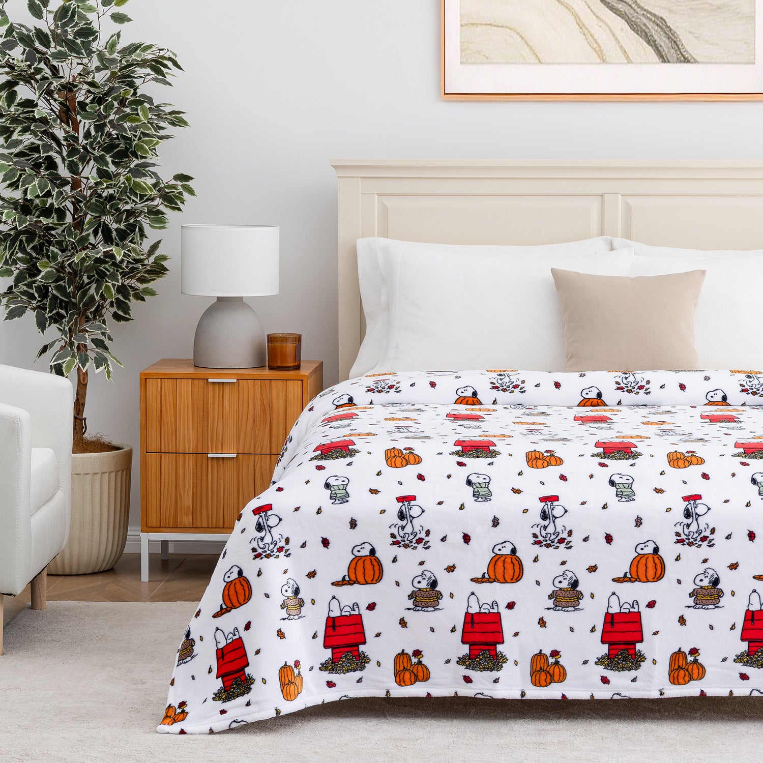 A bed dressed in a white comforter or sheet set printed with a repeating cartoon pattern of Snoopy and his doghouse, pumpkins, leaves, and autumn motifs, styled in a bright bedroom with a wooden nightstand, lamp, plant, and white headboard.