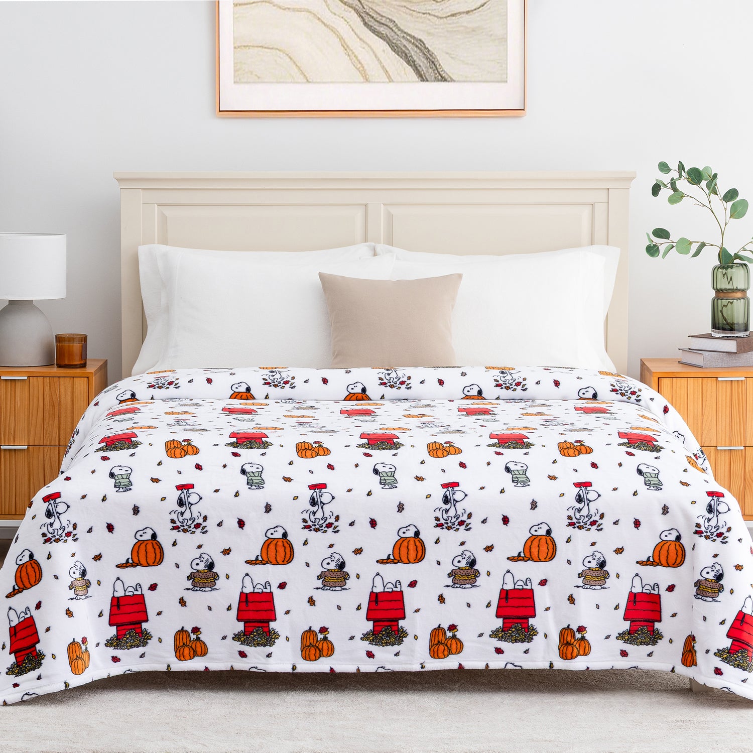 A white bedding set, including a comforter and matching pillow shams, featuring a repeating cartoon pattern of Snoopy and his doghouse, pumpkins, and leaves. The bed is styled in a bright, modern bedroom with a light wood headboard, bedside tables, and a framed picture on the wall.