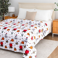 A bed dressed in a white comforter set featuring a repeating cartoon print of Snoopy and Woodstock in red, orange, black, and gray, styled in a bright bedroom with white pillows, a beige accent pillow, wooden nightstands, and a lamp.