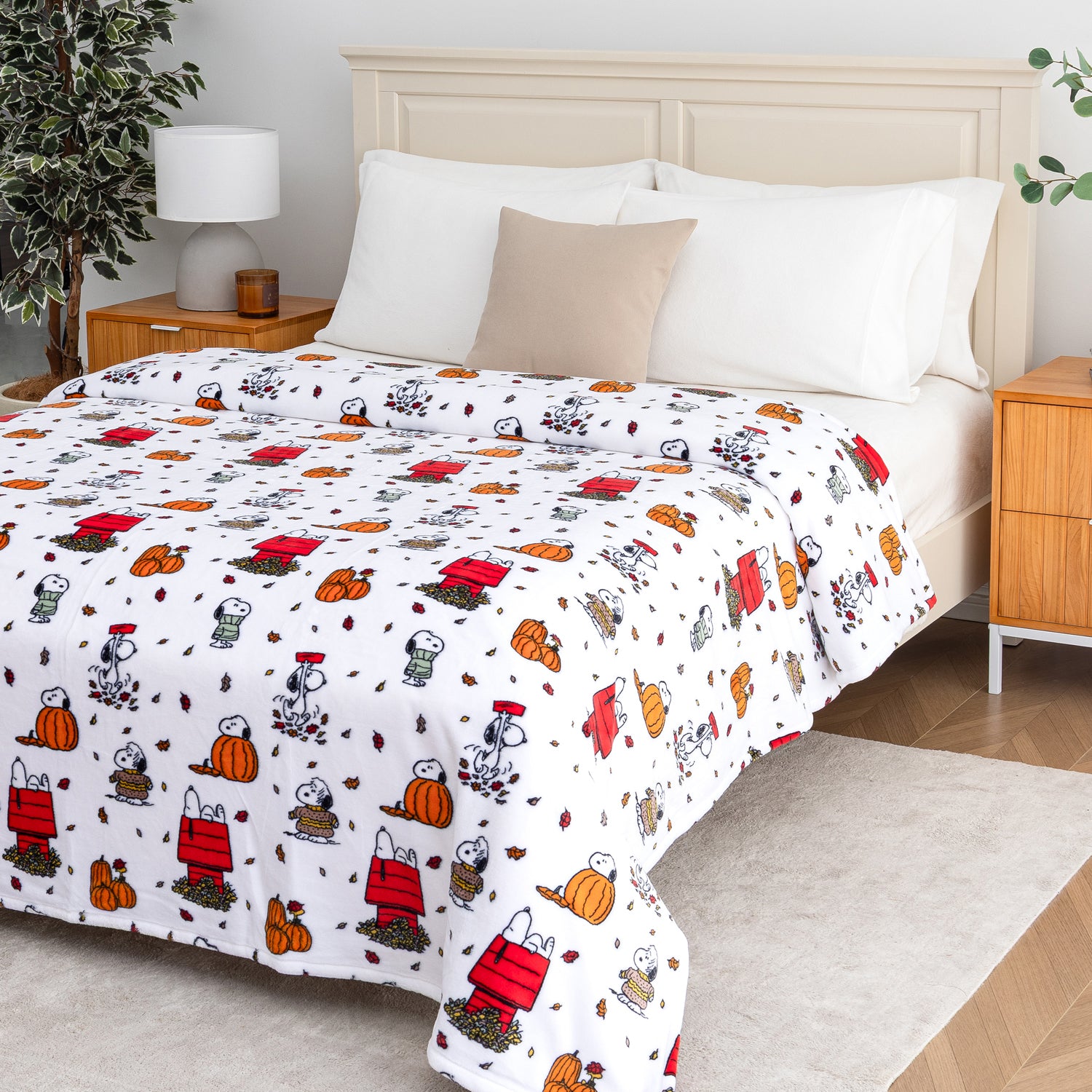A bed dressed in a white comforter set featuring a repeating cartoon print of Snoopy and Woodstock in red, orange, black, and gray, styled in a bright bedroom with white pillows, a beige accent pillow, wooden nightstands, and a lamp.
