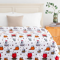 A white bedding set with a repeating cartoon print of Snoopy and Woodstock in autumn outfits, styled on a bed in a bedroom setting with a wooden headboard, beige pillow, and bedside table.