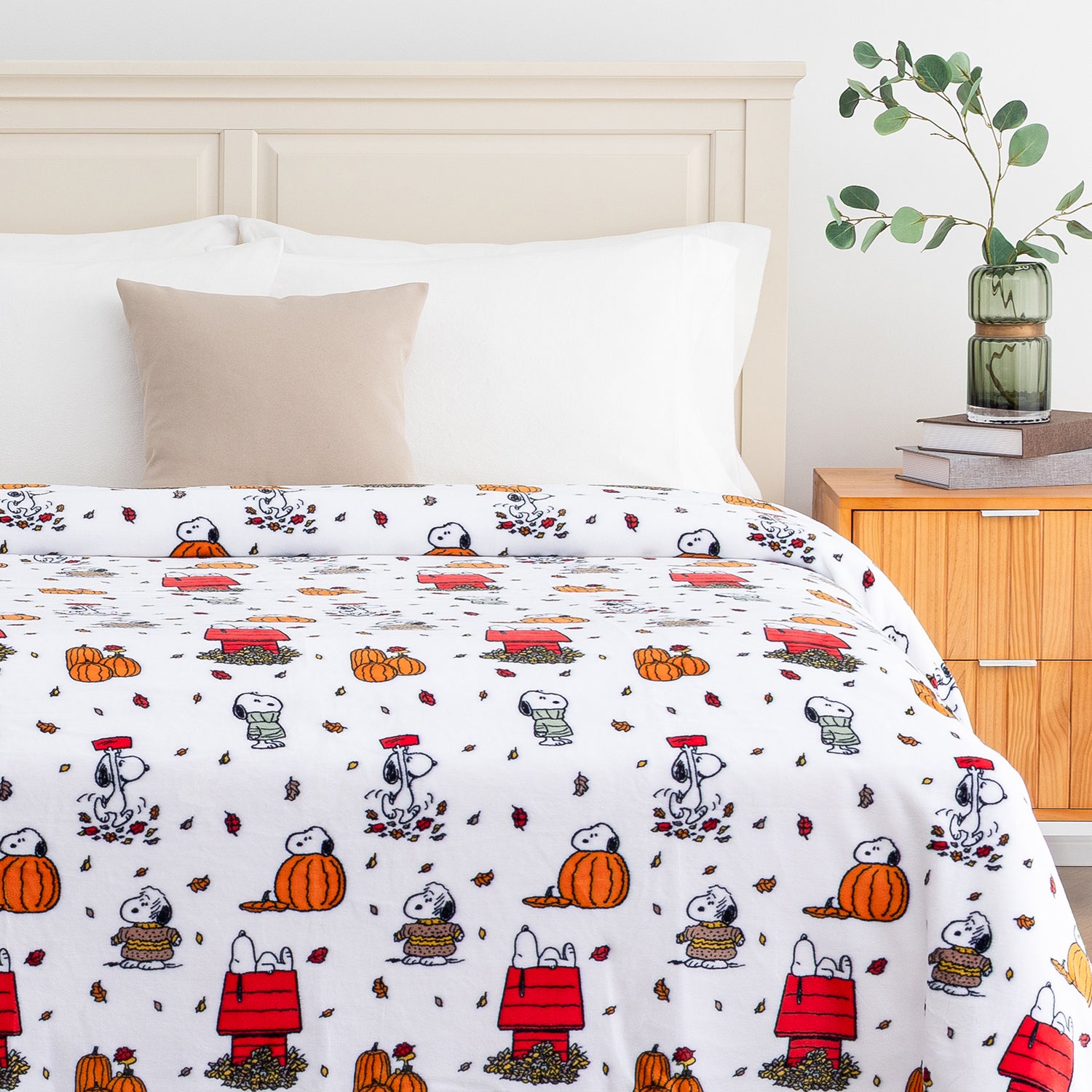 A white bedding set with a repeating cartoon print of Snoopy and Woodstock in autumn outfits, styled on a bed in a bedroom setting with a wooden headboard, beige pillow, and bedside table.