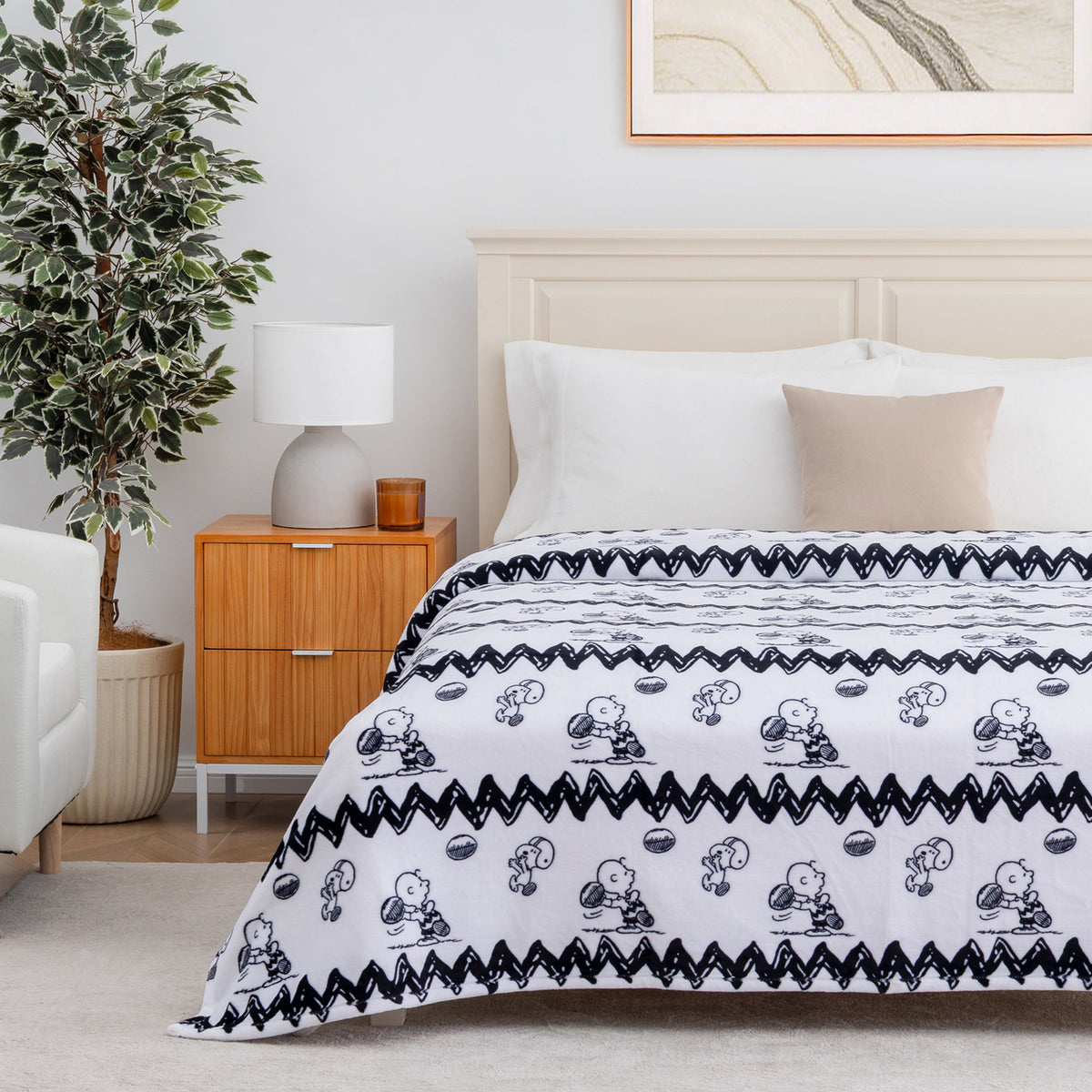 A white blanket with a black Snoopy and Woodstock pattern and zigzag border, styled on a bed in a bright, modern bedroom with a wooden nightstand, potted plant, and white chair.