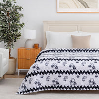 A white blanket with a black Snoopy and Woodstock pattern and zigzag border, styled on a bed in a bright, modern bedroom with a wooden nightstand, potted plant, and white chair.