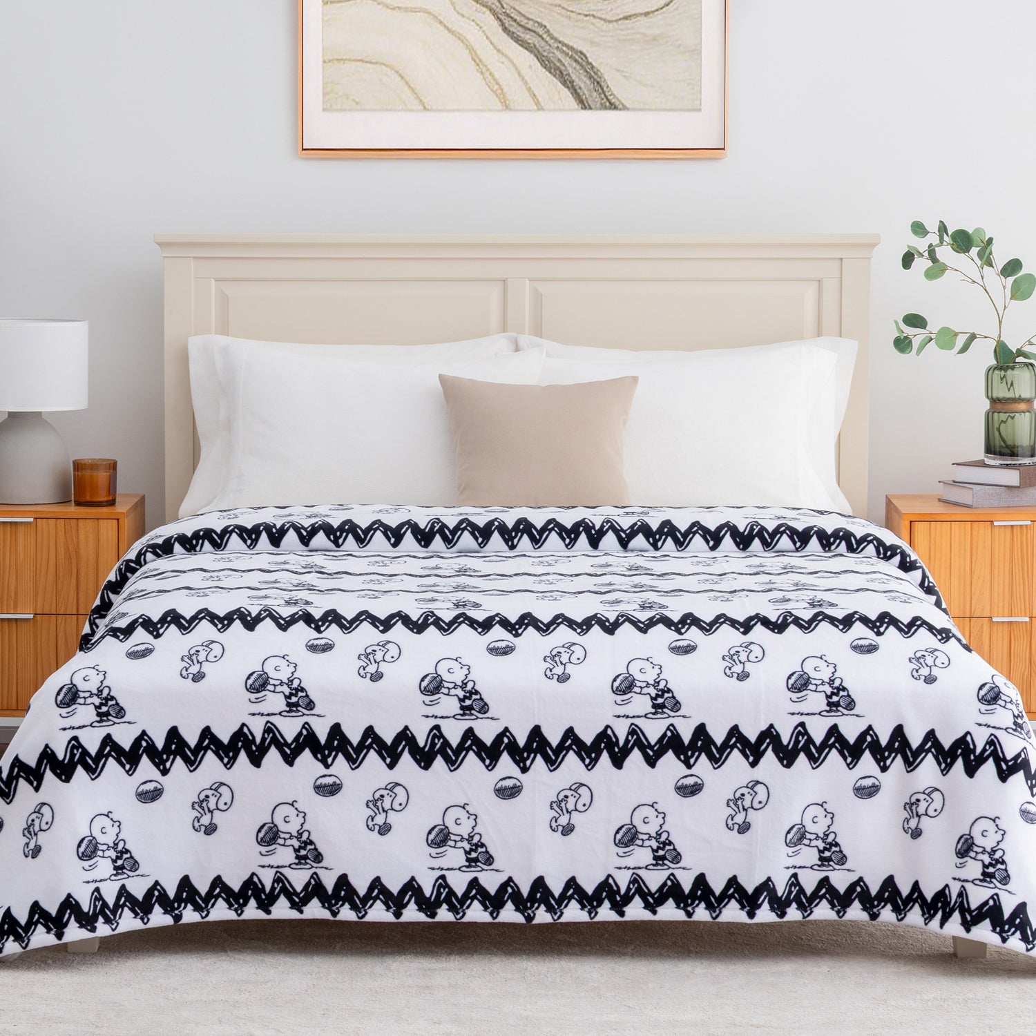 A bed dressed in a white comforter or duvet with a black zigzag border and repeated black cartoon character prints, styled in a bright bedroom with white pillows, a beige accent pillow, wooden nightstands, a lamp, and framed wall art.