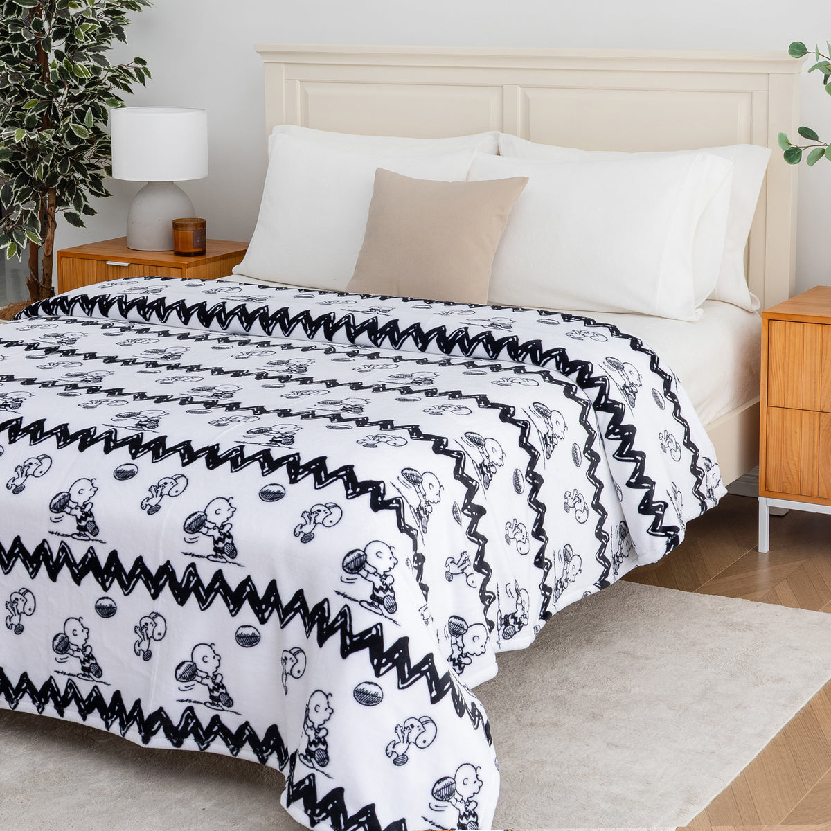 A white blanket with black zigzag borders and black-and-white cartoon character prints is styled on a bed in a bright, modern bedroom setting.