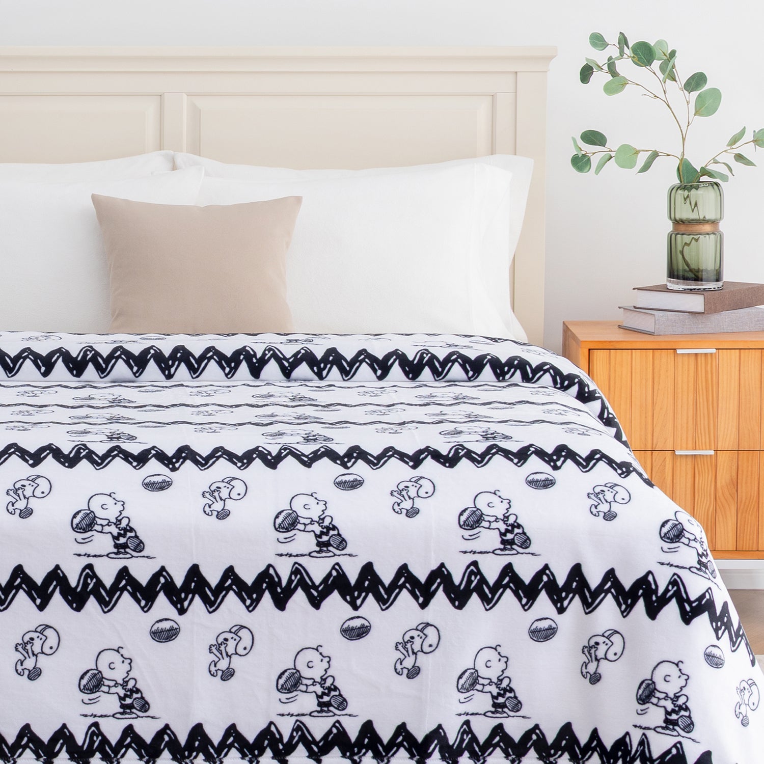 A white blanket with a black Snoopy and Woodstock pattern and zigzag border, styled on a bed in a bright, modern bedroom with a wooden nightstand, potted plant, and white chair.