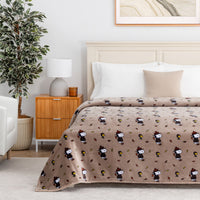 A beige blanket with a repeating cartoon panda print is styled on a bed in a bright, modern bedroom setting with a wooden nightstand, lamp, and potted plant.