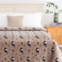 A cozy bed setup featuring a tan blanket and matching pillow cover with a repeating pattern of Snoopy as a Thanksgiving turkey wearing a hat, styled with white pillows and a beige accent pillow in a bedroom setting.