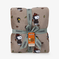 A folded plush blanket with a repeating Peanuts character print on a warm taupe background, wrapped with a gray ribbon and an orange Peanuts tag.