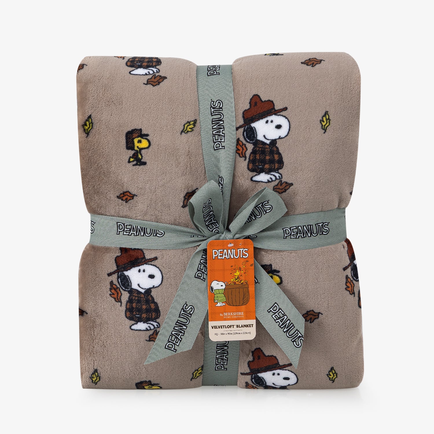 A folded plush blanket with a repeating Peanuts character print on a warm taupe background, wrapped with a gray ribbon and an orange Peanuts tag.