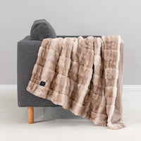 A plush faux-fur throw blanket in a warm taupe and beige patchwork pattern draped over a modern gray armchair with a wooden leg, set against a neutral light gray background.