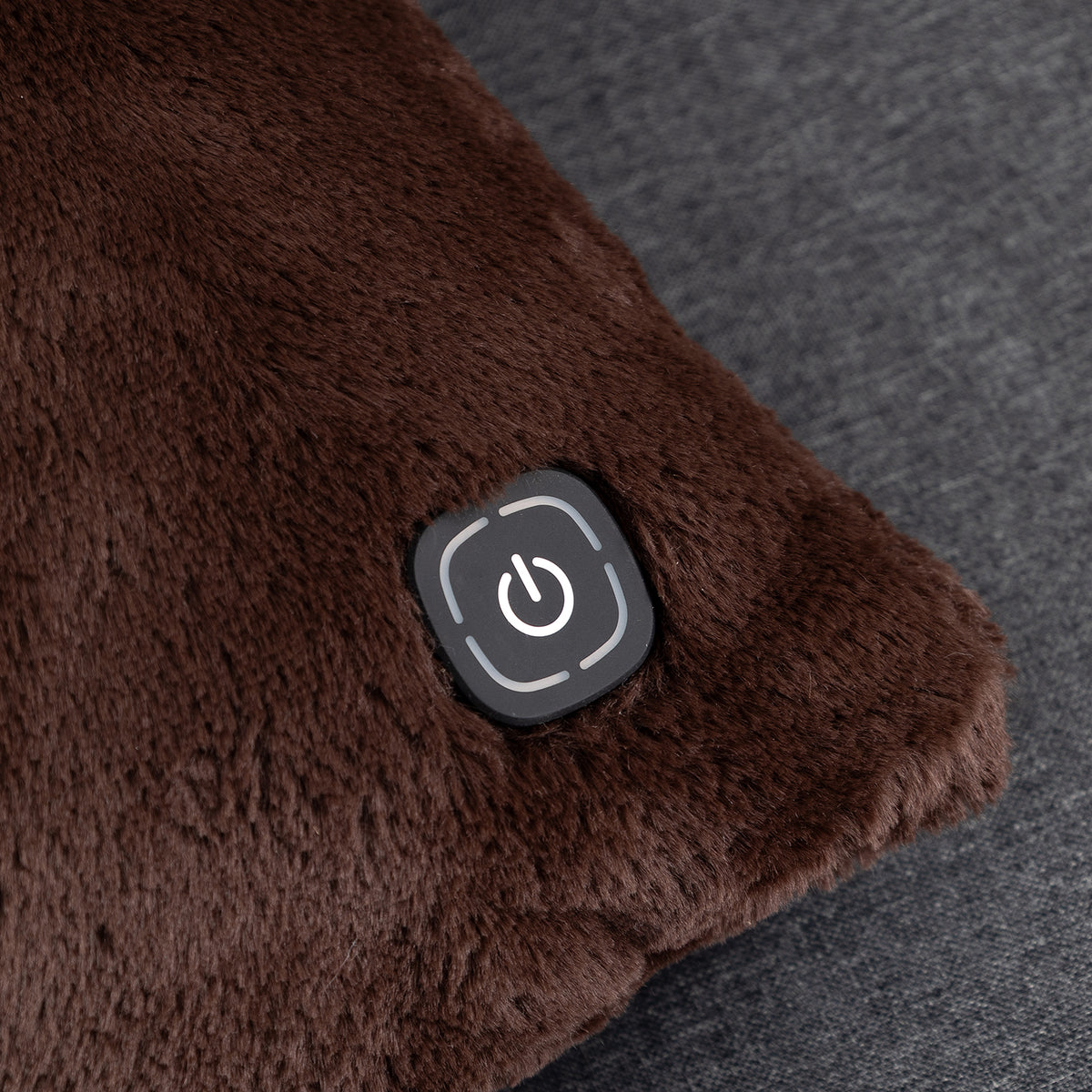 Close-up of a brown plush heated cushion with a black power button on a dark gray textured fabric background.