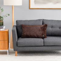 A dark brown plush throw pillow with a small circular control button, resting on a gray fabric sofa in a modern living room setting.