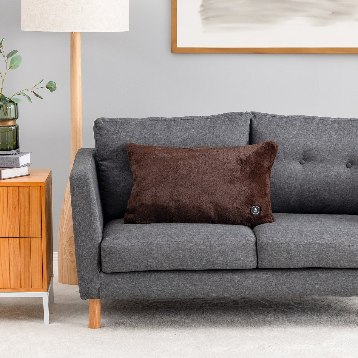 A dark brown plush throw pillow with a small circular control button, resting on a gray fabric sofa in a modern living room setting.