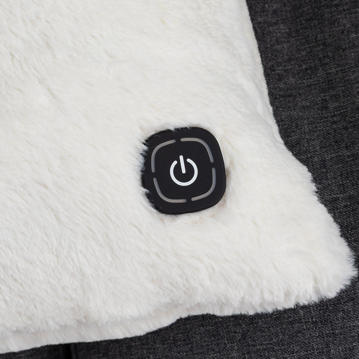 Close-up of a white sherpa or faux-fur cushion with a black square power button on the front, set against a dark gray textured fabric background.