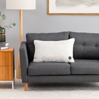 A white fluffy throw pillow with a small black control button on the corner, resting on a dark gray fabric sofa in a modern living room setting.
