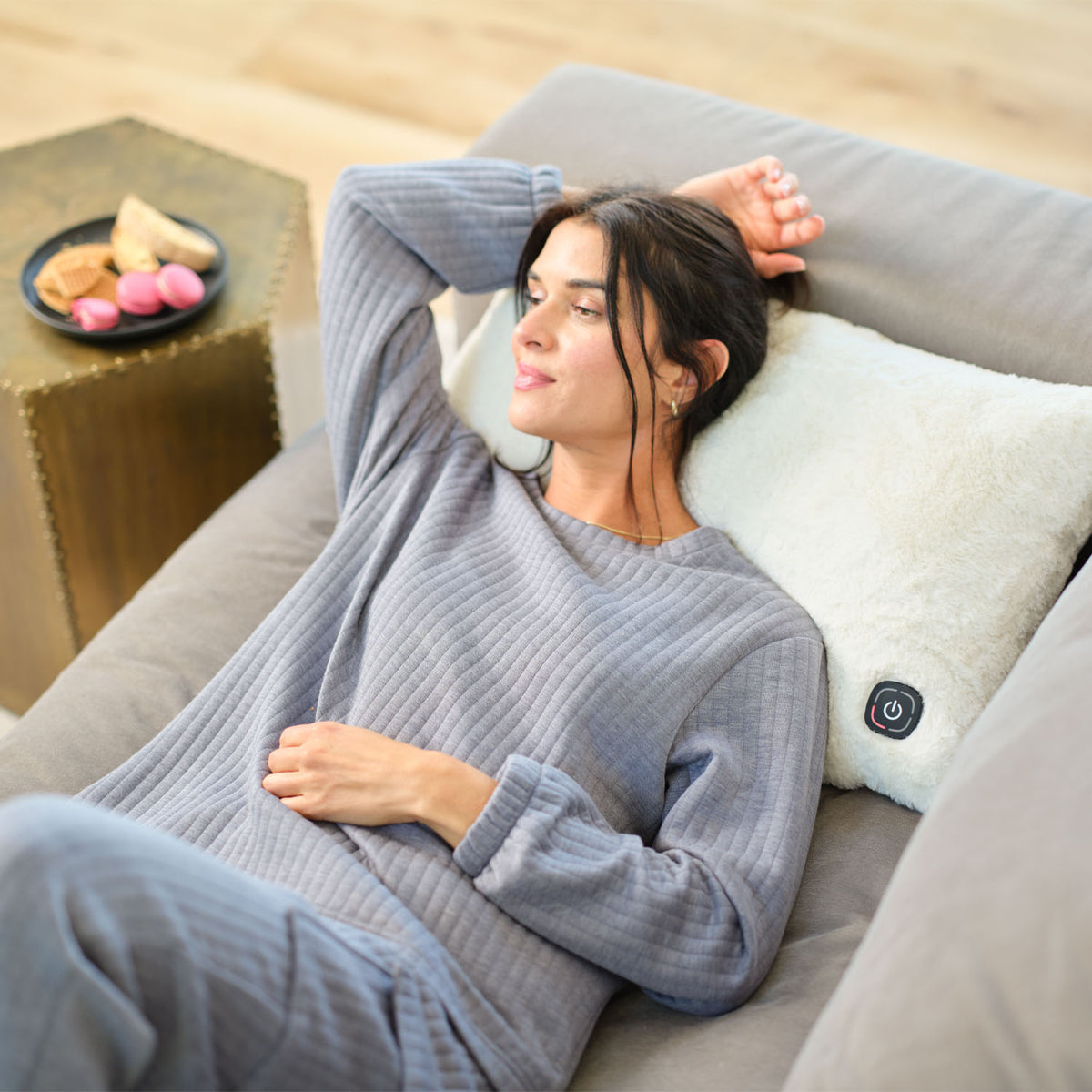 A woman laying on a sofa with a cream heated faux fur lumbar pillow.
