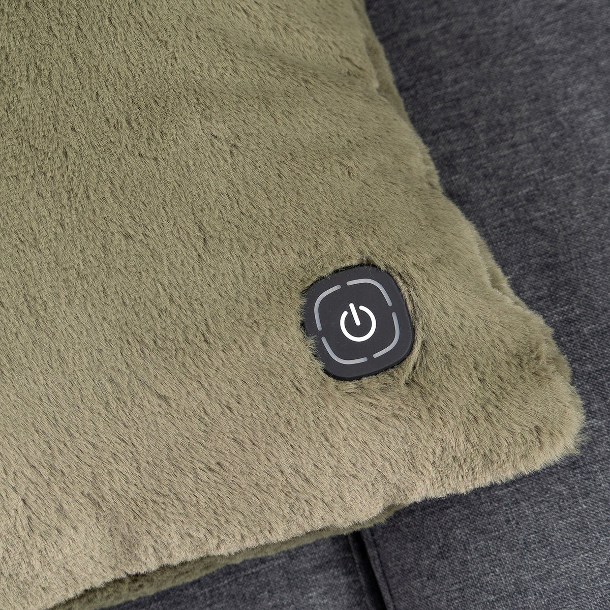 Close-up of a plush, textured electric heated throw pillow in olive green, featuring a black square control button with a power symbol on the corner, resting on a dark gray fabric surface.