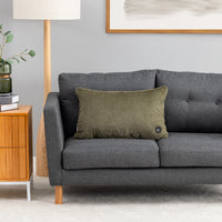 An olive green textured throw pillow with a small black button control on the corner, resting on a dark gray fabric sofa in a modern living room setting.