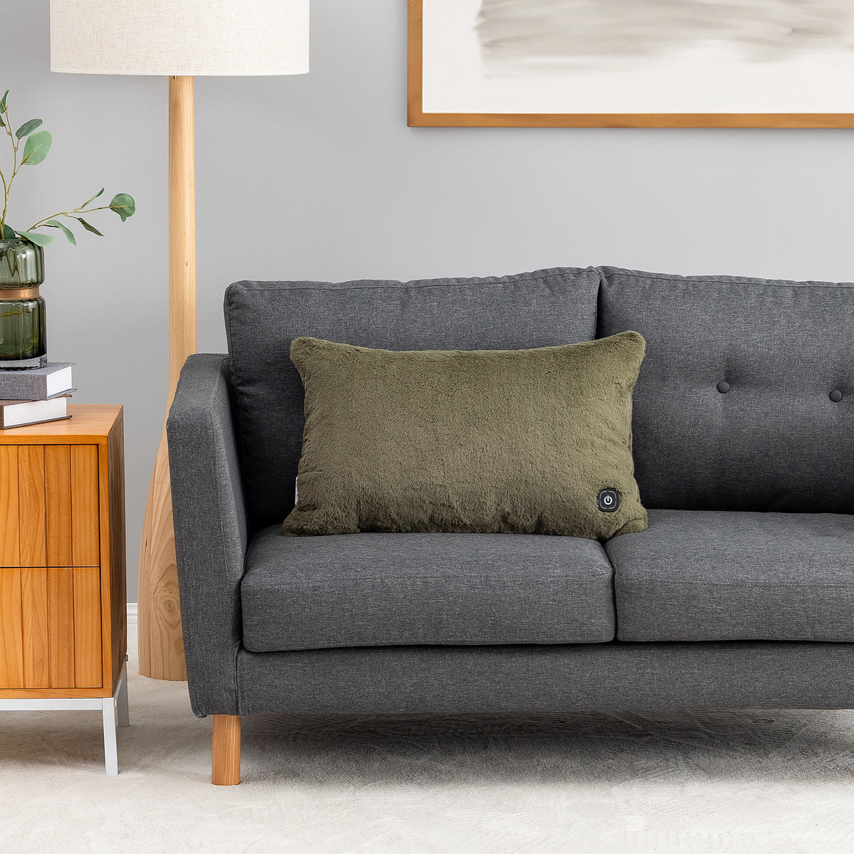 An olive green textured throw pillow with a small black button control on the corner, resting on a dark gray fabric sofa in a modern living room setting.