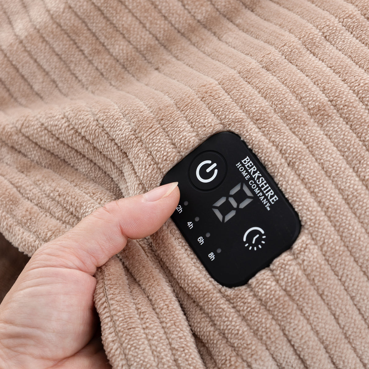 Close-up of a beige ribbed fabric blanket with a black digital control patch held by a hand. The patch shows the brand name BERKSHIRE HOME COMPANY and control icons.