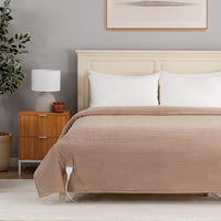 A bed dressed in a ribbed taupe-brown quilt or comforter, styled in a modern bedroom with a cream headboard, white pillows, a wooden nightstand, and a gray wall.