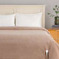 A beige ribbed blanket or duvet cover with a white control cord visible at the corner, styled on a bed with white pillows and a cream headboard in a bedroom setting.