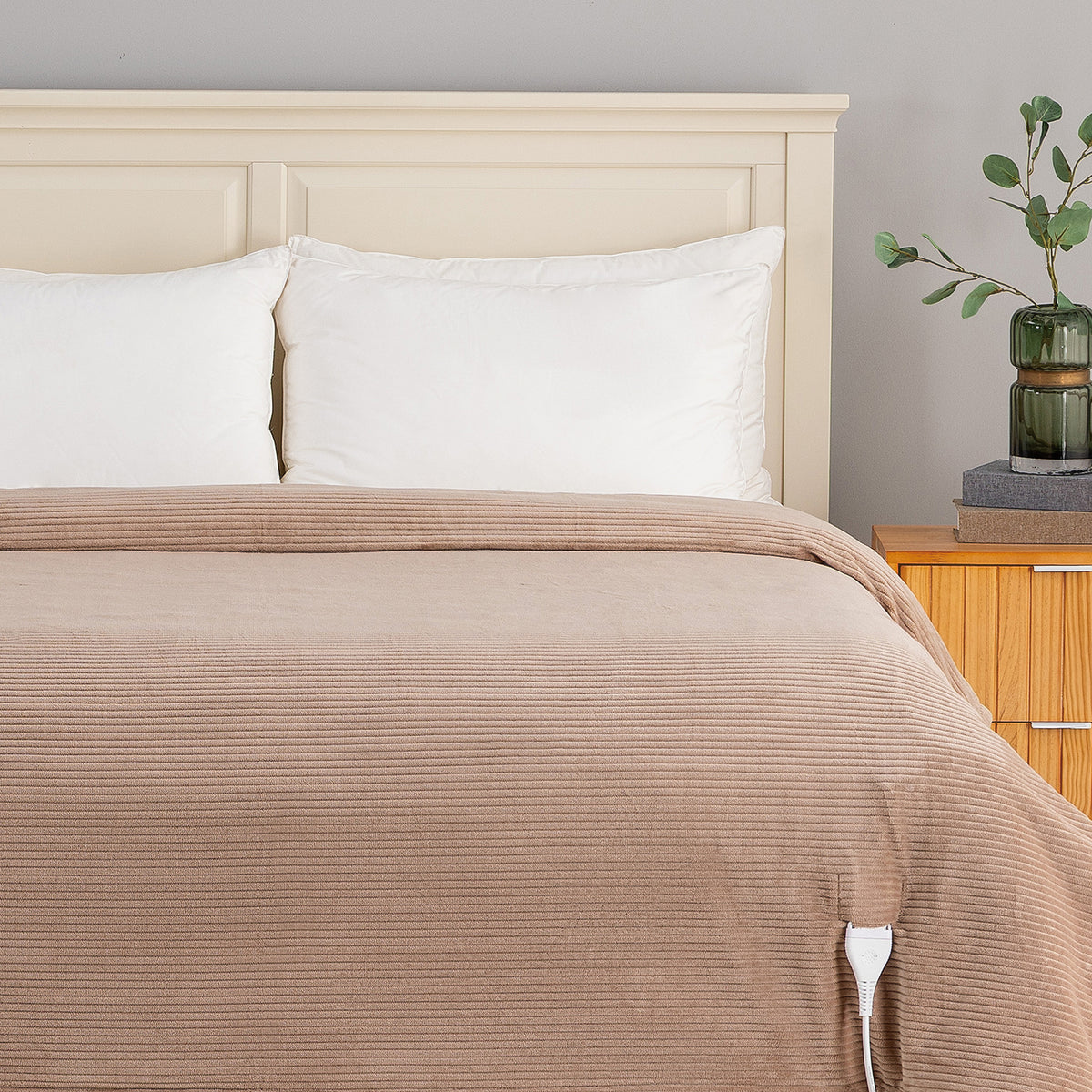 A beige ribbed blanket or duvet cover with a white control cord visible at the corner, styled on a bed with white pillows and a cream headboard in a bedroom setting.