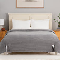 A gray ribbed electric heated blanket with white cord connectors is shown draped over a bed with white pillows and a cream-colored wooden headboard in a bedroom setting.