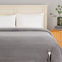 A gray ribbed blanket or comforter spread across a bed with white pillows and a light wood headboard, shown in a styled bedroom setting with a bedside table and vase.