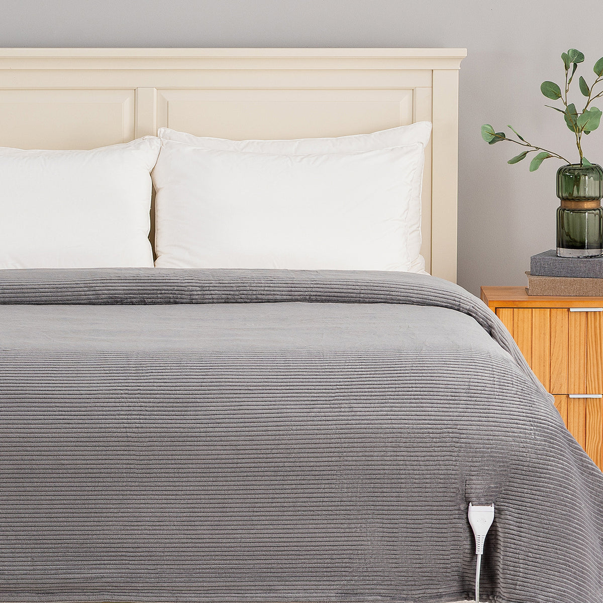 A gray ribbed blanket or comforter spread across a bed with white pillows and a light wood headboard, shown in a styled bedroom setting with a bedside table and vase.