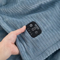 A close-up of a blue ribbed blanket with an integrated black digital control pad being held by a hand, showing touch controls and a display screen.
