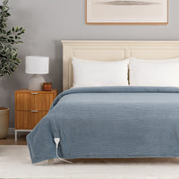 A blue ribbed blanket with an integrated white heating pad is shown on a bed in a modern bedroom setting. The scene includes a wooden nightstand with a lamp, a potted plant, and a light gray wall.