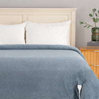 A close-up of a bed dressed with a slate blue corduroy comforter and white pillows, set against a light beige wooden headboard with a wooden nightstand and a green glass vase on a gray wall.