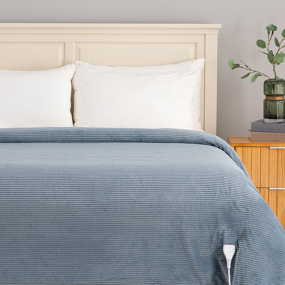 A close-up of a bed dressed with a slate blue corduroy comforter and white pillows, set against a light beige wooden headboard with a wooden nightstand and a green glass vase on a gray wall.