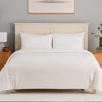 A bed dressed in a cream-colored ribbed duvet cover and matching pillowcases, set against a light wood headboard in a styled bedroom with bedside tables, a lamp, and wall art.