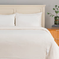 A bed dressed in white ribbed bedding, including a duvet cover and two pillows, set against a light wood headboard. A wooden nightstand with a green glass vase is visible on the right against a neutral gray wall.